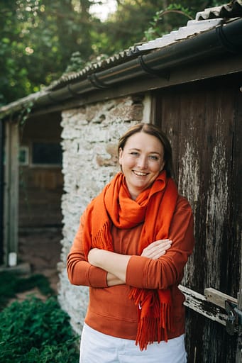 Smiling woman in orange scarf and orange top, standing in front of a rustic farm building